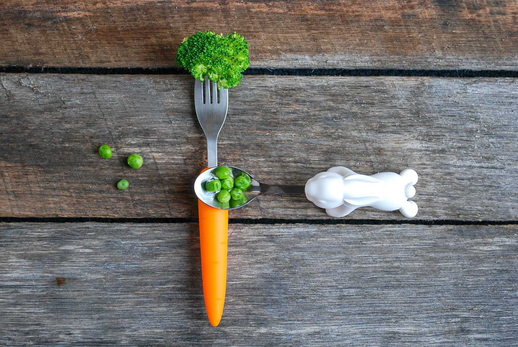 A fork with a handle like a carrot holds a piece of broccoli, and a spoon with a handle shaped like a rabbit holds some peas. Both are sitting on a wood table.