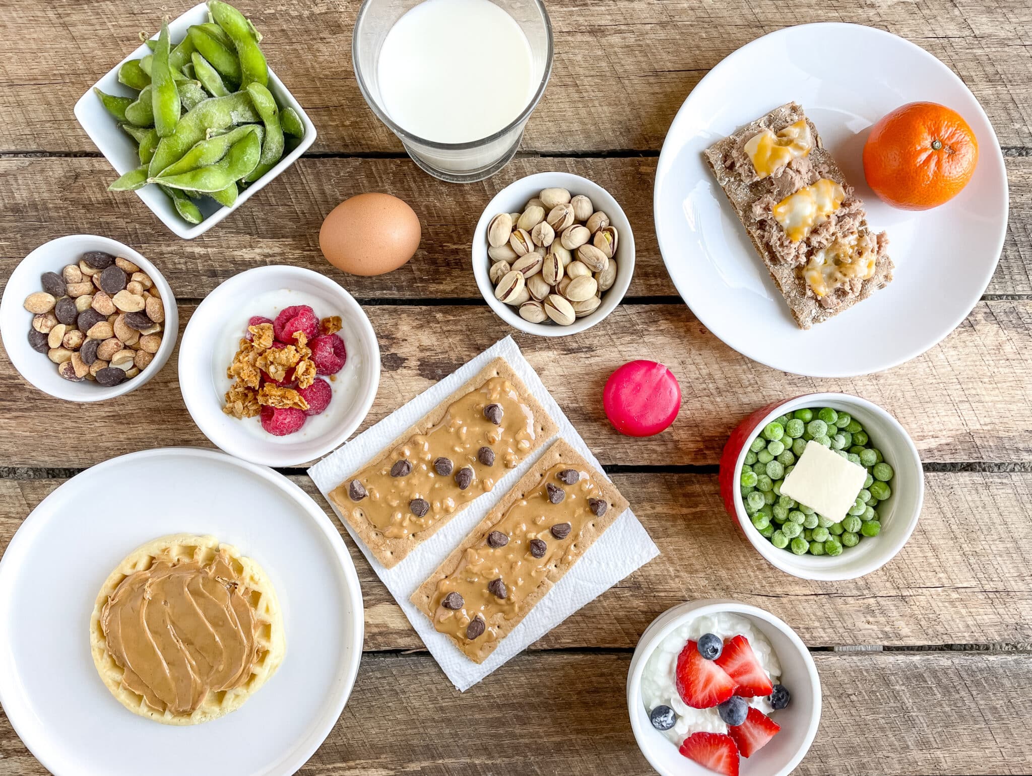 A variety of protein snacks for kids sit on a wood table, including edamame, yogurt and fruit, cheese, hard-boiled egg, milk, and green peas.