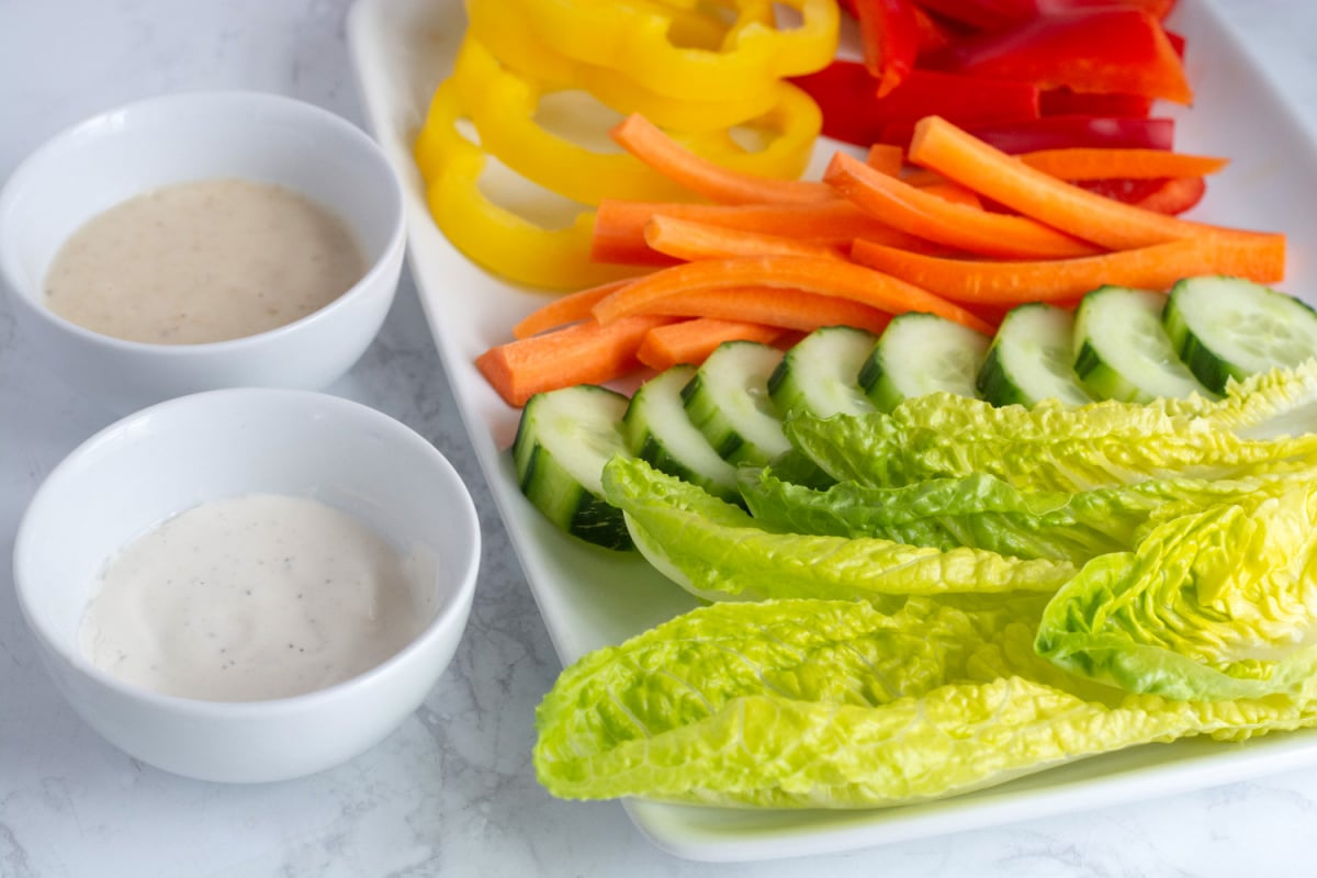 A kids salad is deconstructed on a plate: Lettuce leaves, cucumbers, carrots, and peppers next to small dishes of dressing.