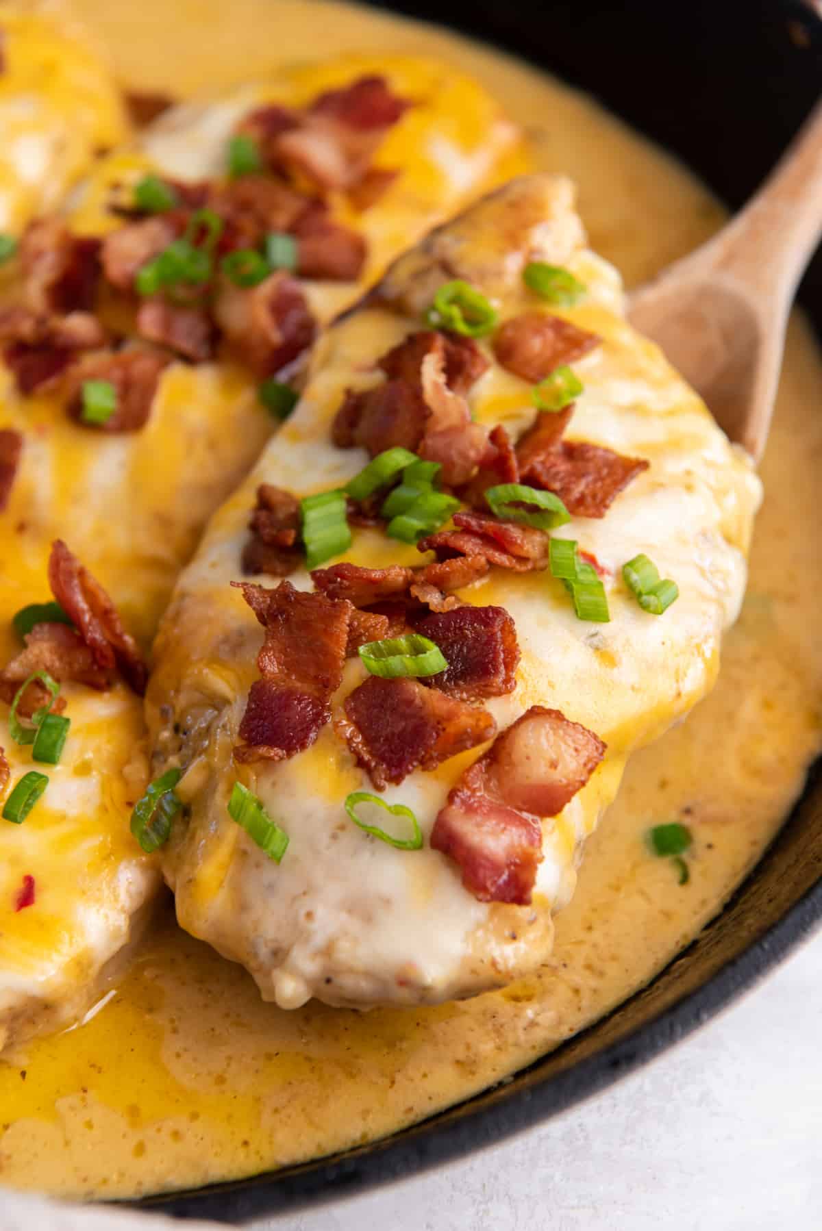 a wooden spoon lifting a cheesy bacon ranch chicken breast from a cast iron skillet.