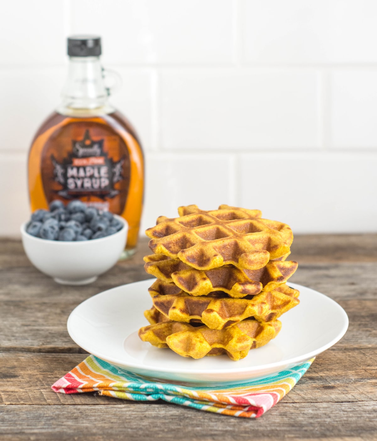 A white plate with four Pumpkin Protein Waffles sits on a wood table with a jar of maple syrup and bowl of blueberries in the background.