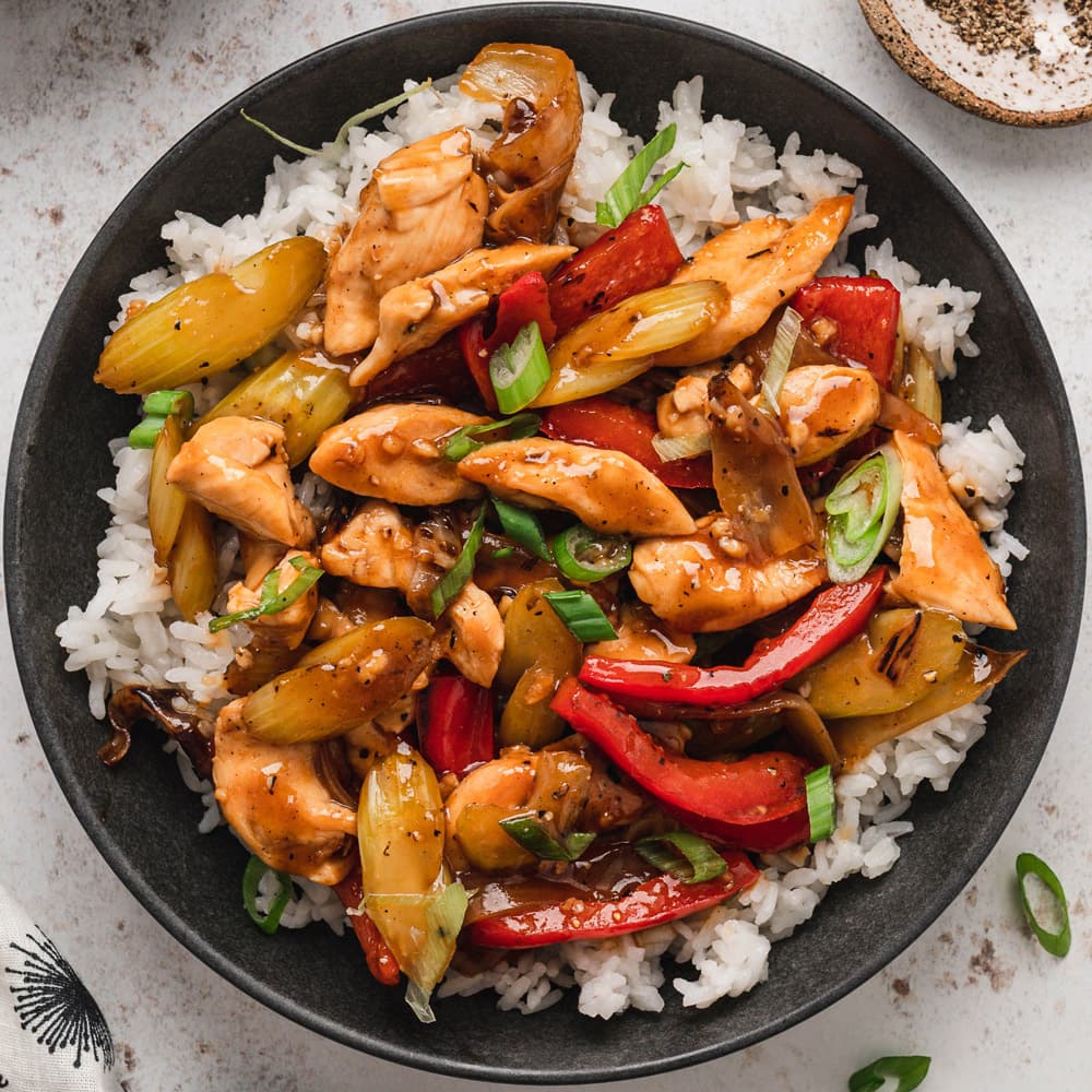 Chicken and vegetables in a brown sauce over steamed rice on a black plate.